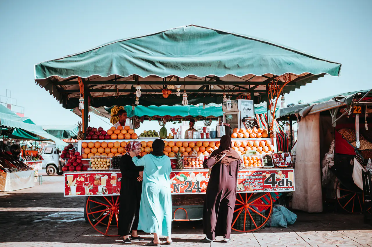 Women at a fresh orange juice stall in a market square.